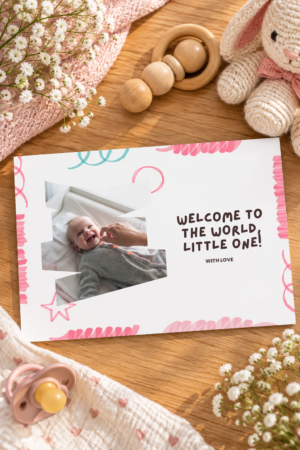 A cute white greeting card featuring a jagged photo cutout of a smiling baby in a grey onesie, next to playful text that reads Welcome to the world, little one! with pink and blue doodles.