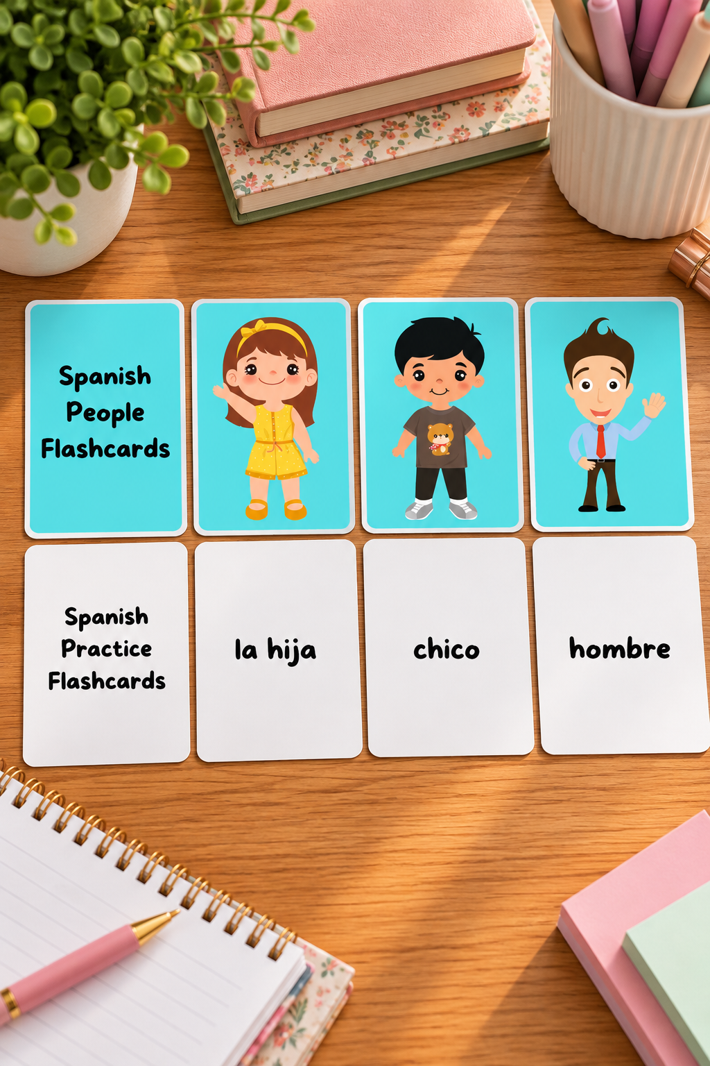 A top down photo of eight educational flashcards laid out horizontally on a smooth wooden table with school notebooks and a pen. The top row features bright cyan blue cards showing cartoon characters of a waving girl in a yellow dress, a boy in a teddy bear t-shirt, and a smiling man in a tie, while the matching bottom row consists of white practice cards with the Spanish words la hija, chico, and hombre.